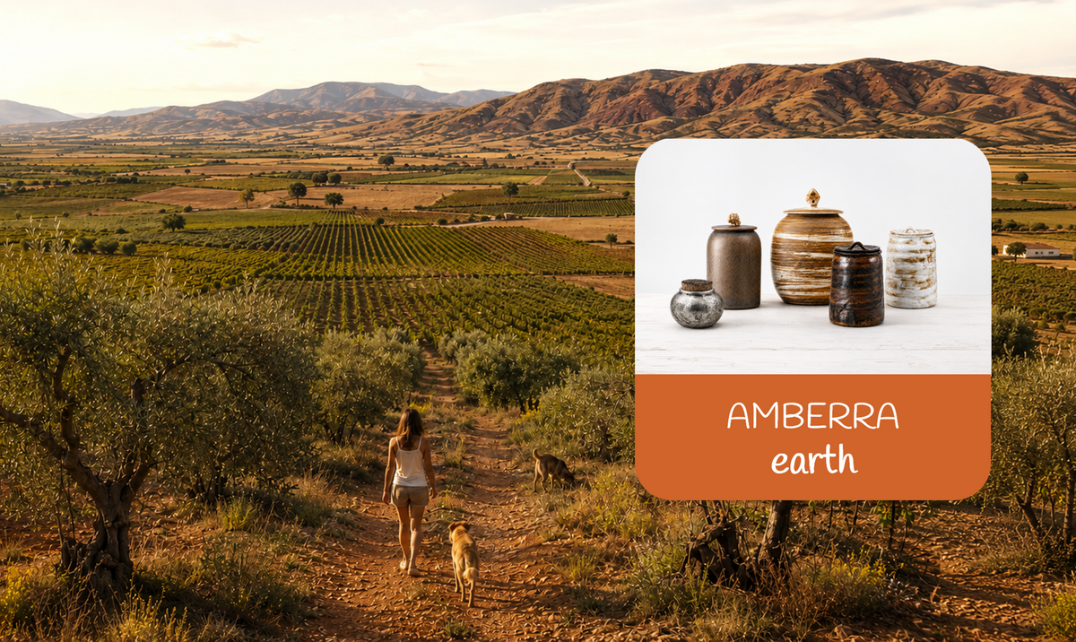 Outdoor landscape scene with a woman walking a dirt path through vineyards and hills, with a product inset showing ceramic urns AMBERRA earth.