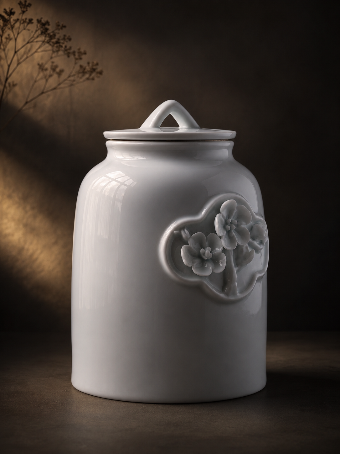 A glossy white ceramic jar with a matching lid and a raised floral relief decoration on the front, photographed against a dark brown background with warm light and dried branches.